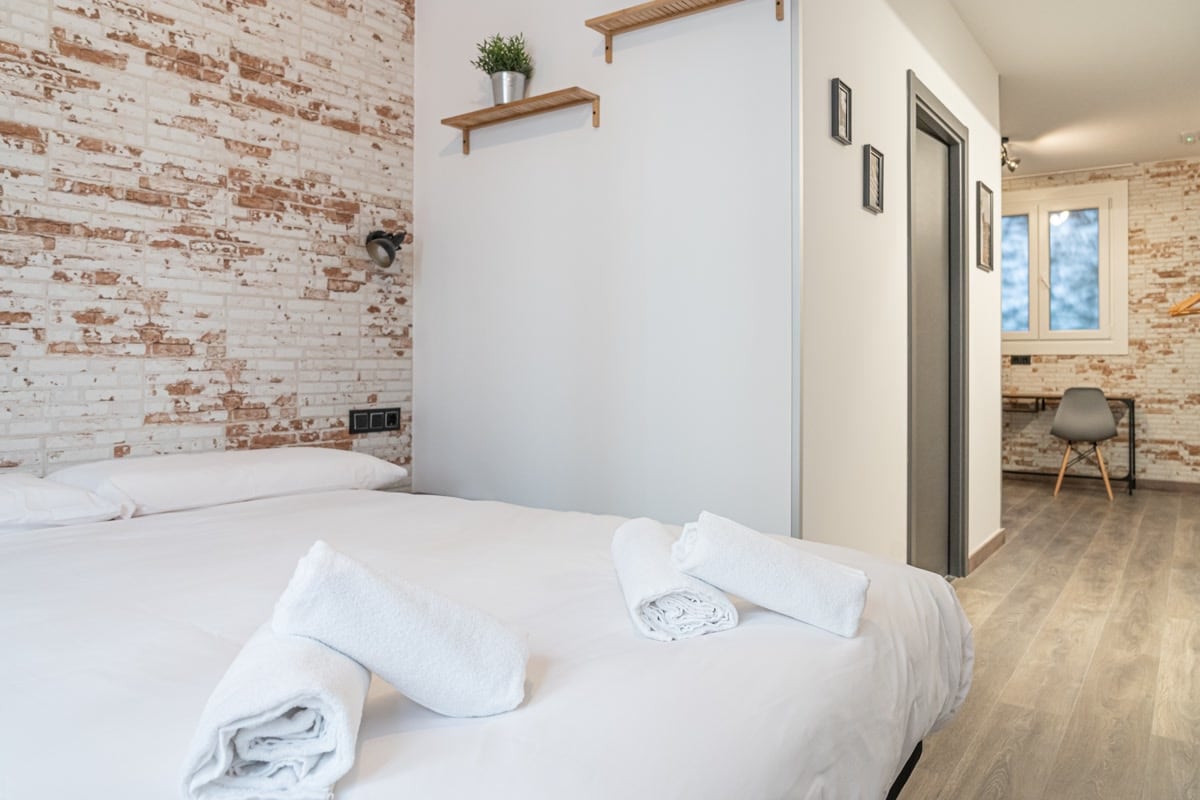 A spacious sleeping area features a comfortable bed with neatly arranged linen. Soft, white towels are placed on the bed and a small shelf with a decorative plant can be seen on the exposed brick wall. A workspace with a chair is positioned nearby, illuminated by natural light from a window.
