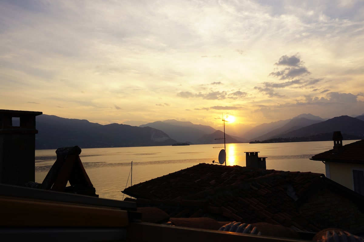 A scenic view features a sunset over a tranquil lake, with mountains in the background. The sun casts a golden reflection on the water, while rooftops of nearby buildings are visible in the foreground. Soft clouds add texture to the evening sky.
