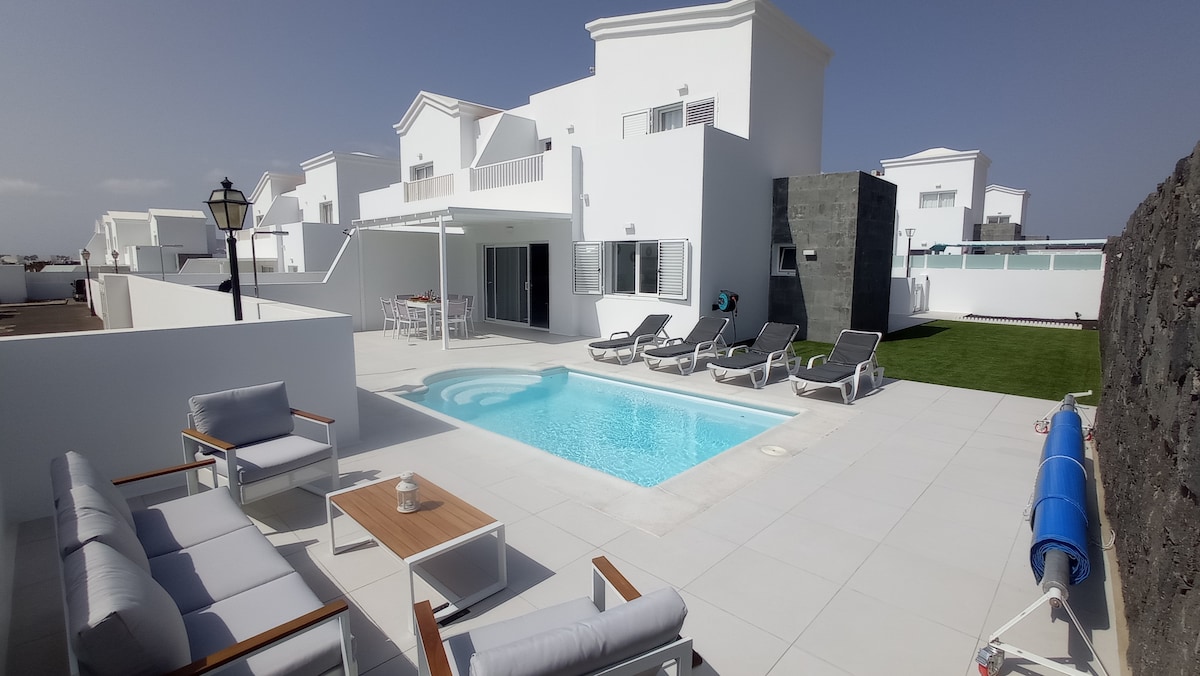 An outdoor area features a private swimming pool surrounded by paved flooring. Comfortable lounge chairs are positioned near the pool, alongside a small seating arrangement with a table. A well-maintained green space and a barbecue area are also visible in this tranquil setting.