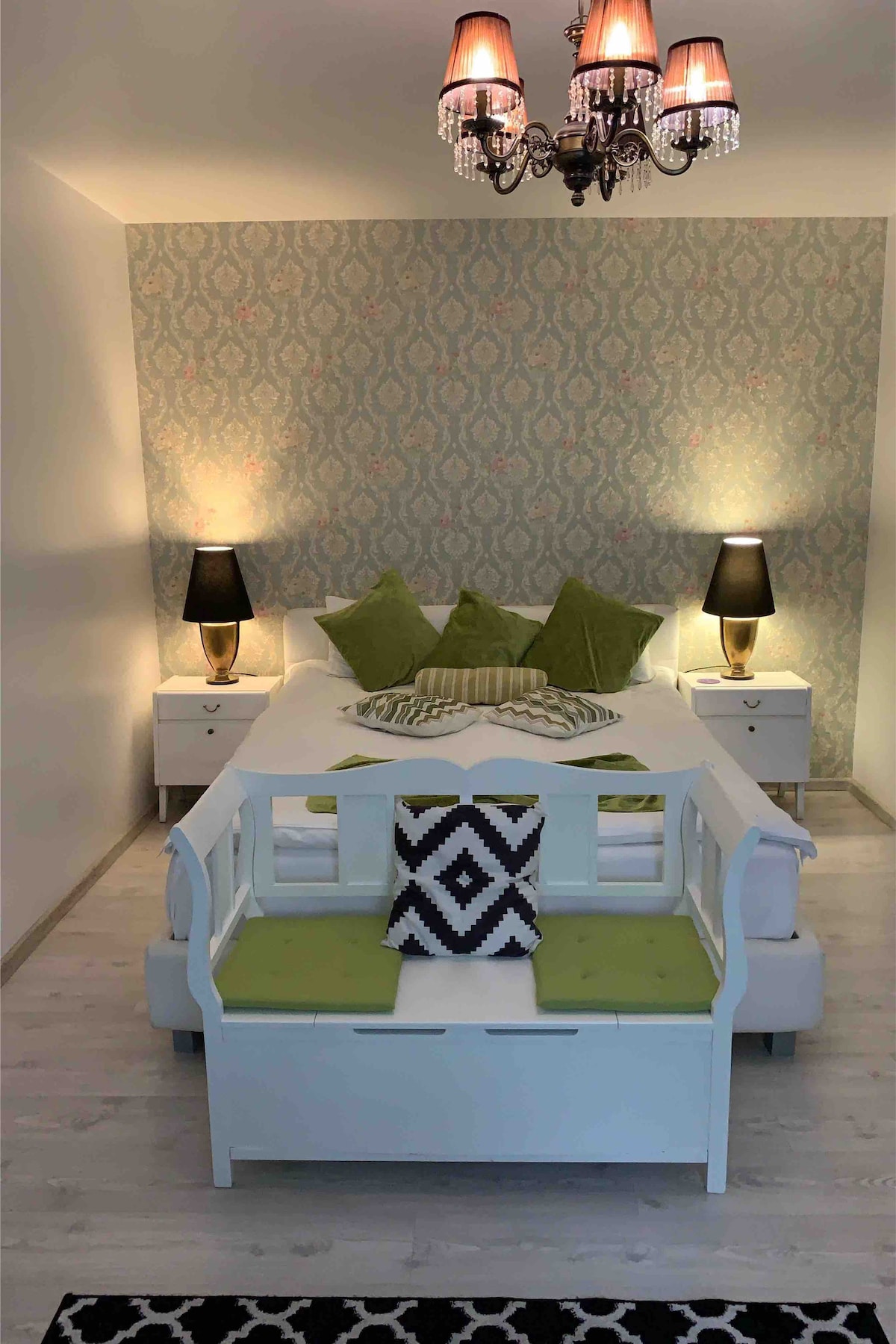 A spacious bedroom features a large bed framed by decorative green cushions. Two bedside tables with lamps accentuate the space, while a stylish bench with a decorative pillow adds seating. A chandelier provides soft, ambient illumination against a patterned wall.
