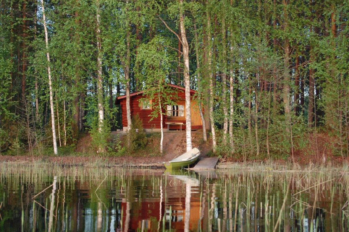 A rustic wooden cottage is nestled among tall trees, reflecting in the calm waters of a nearby lake. A small rowing boat is visible at a dock, adding to the serene lakeside setting surrounded by lush greenery.