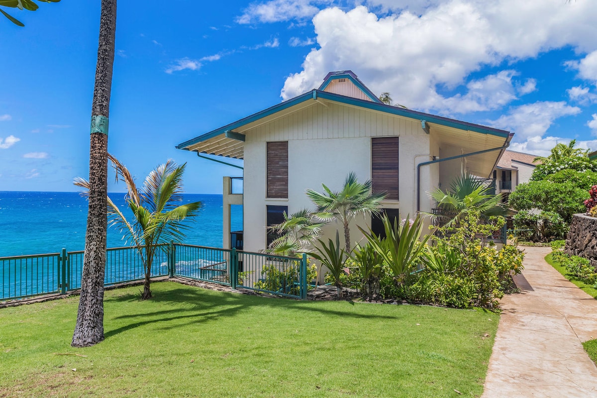 The exterior of a corner unit is seen, surrounded by lush greenery and vibrant tropical plants. The ocean is visible in the background, with blue waters meeting a partly cloudy sky. A pathway leads up to the entrance, creating a welcoming approach.