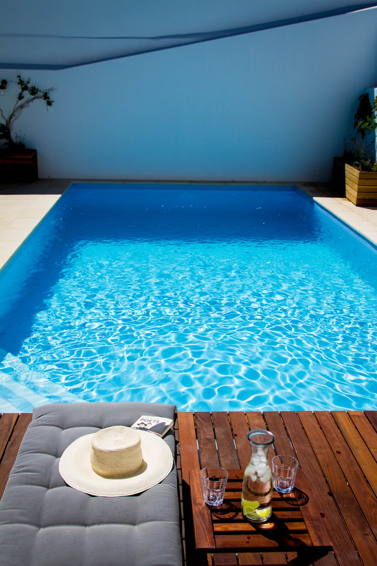 A refreshing saltwater swimming pool is surrounded by smooth tiles and light-colored walls, reflecting sunlight. A sun lounger with a wide-brimmed hat rests beside a wooden table, which holds two glasses and a carafe. The setting provides a serene place for relaxation.