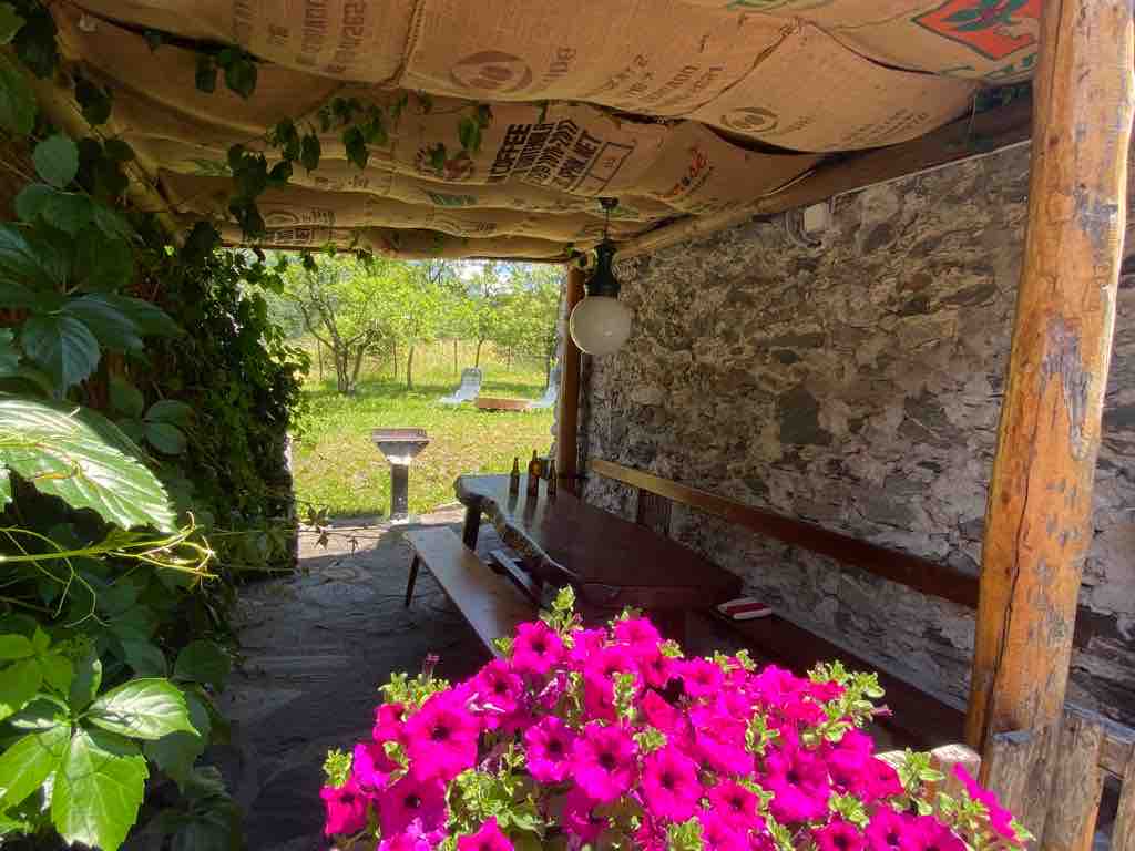 A shaded outdoor seating area features a long wooden table with benches, surrounded by greenery. Bright pink flowers in a pot add color to the scene. The ceiling is covered with natural materials, creating a rustic atmosphere.