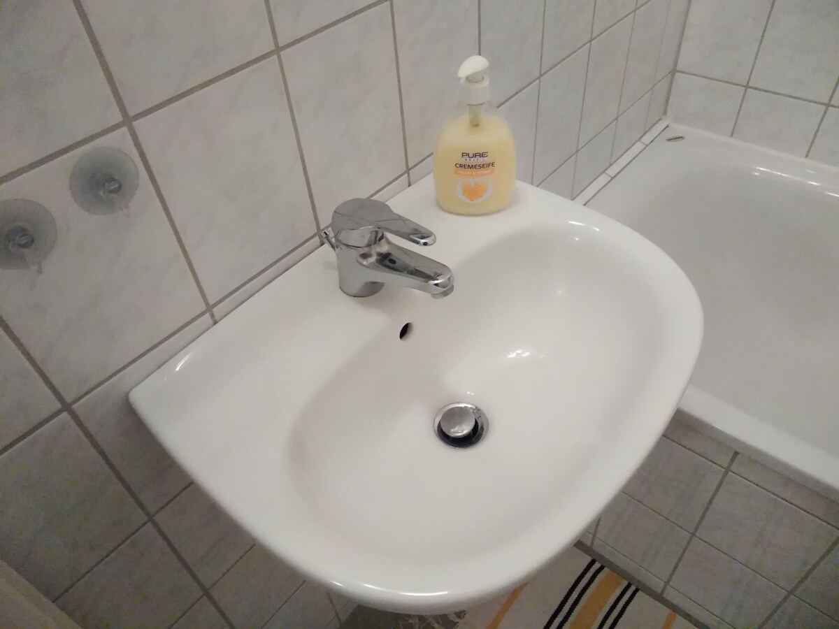 A white bathroom sink is mounted on a tiled wall, featuring a modern faucet. A bottle of liquid soap is placed nearby, and a small drain is visible in the center of the sink. The surrounding tiles create a clean and organized bathroom space.