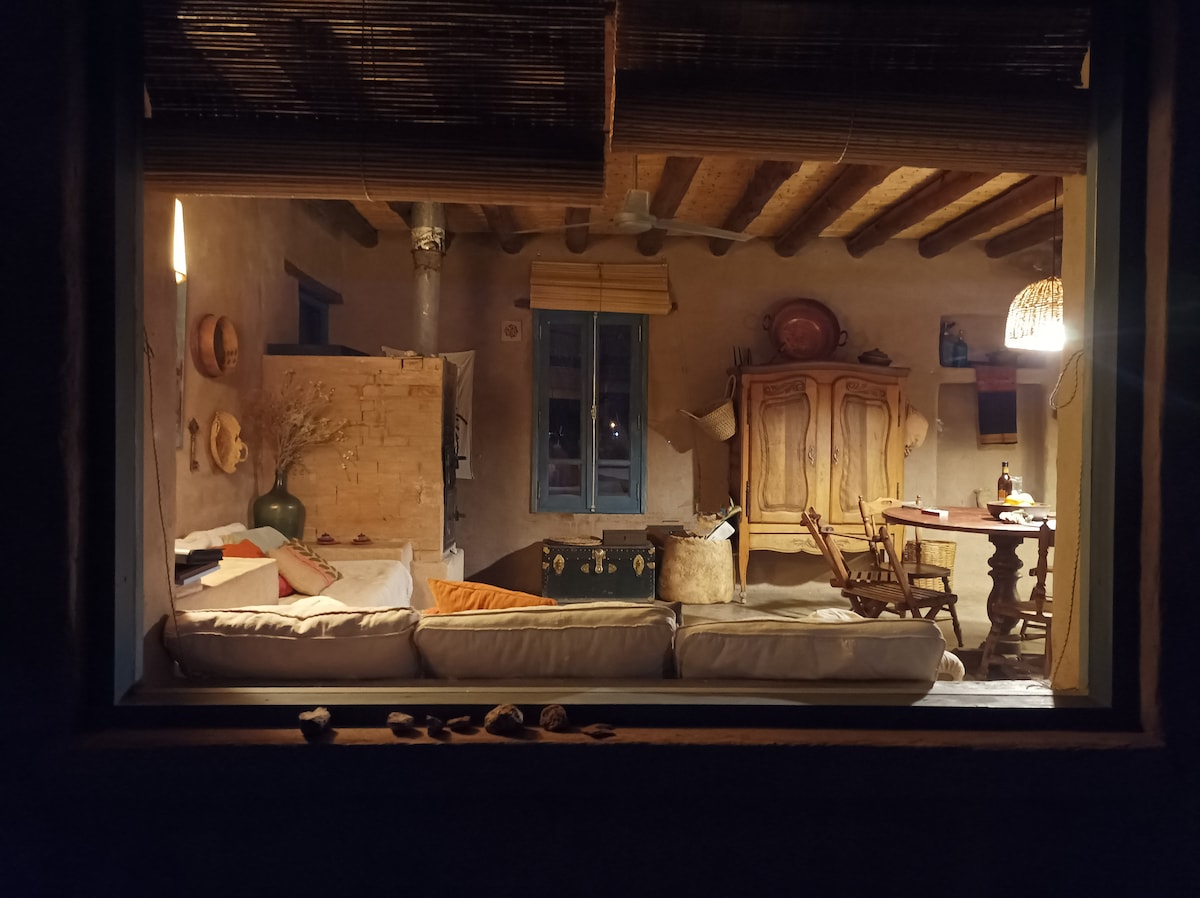 An inviting interior space is visible through a window, featuring a cozy seating area with cushions on a light sofa. A wooden cabinet and a dining table with chairs occupy the room. Soft lighting enhances the warm tones of the adobe walls and exposed wooden beams.