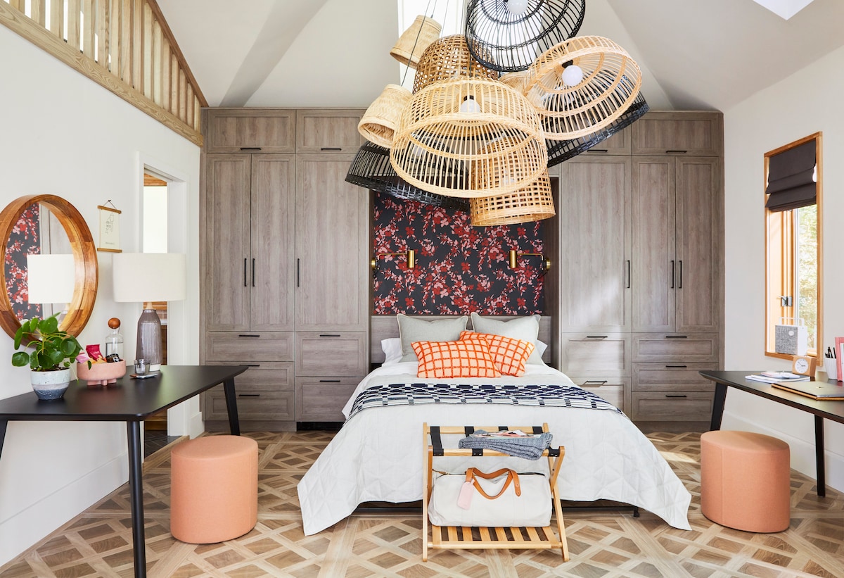 A spacious sleeping area features a central queen bed adorned with a patterned blanket. Two stylish tables are positioned on either side, accompanied by small round stools. An eye-catching display of woven light fixtures hangs above, and contrasting floral wallpaper adds a decorative touch.