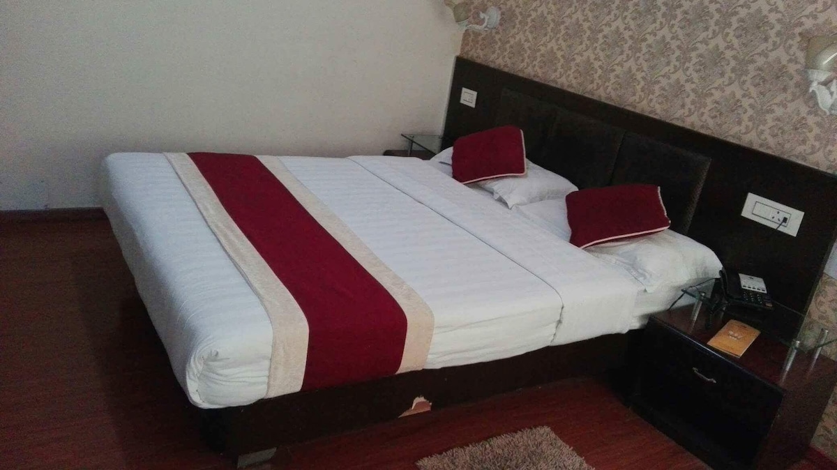 A well-appointed sleeping area features a spacious bed with crisp white linens and coordinating red accent pillows. A decorative rug lies on the wooden floor, while bedside tables with lamps complement the stylish wallpaper in soft neutral tones.