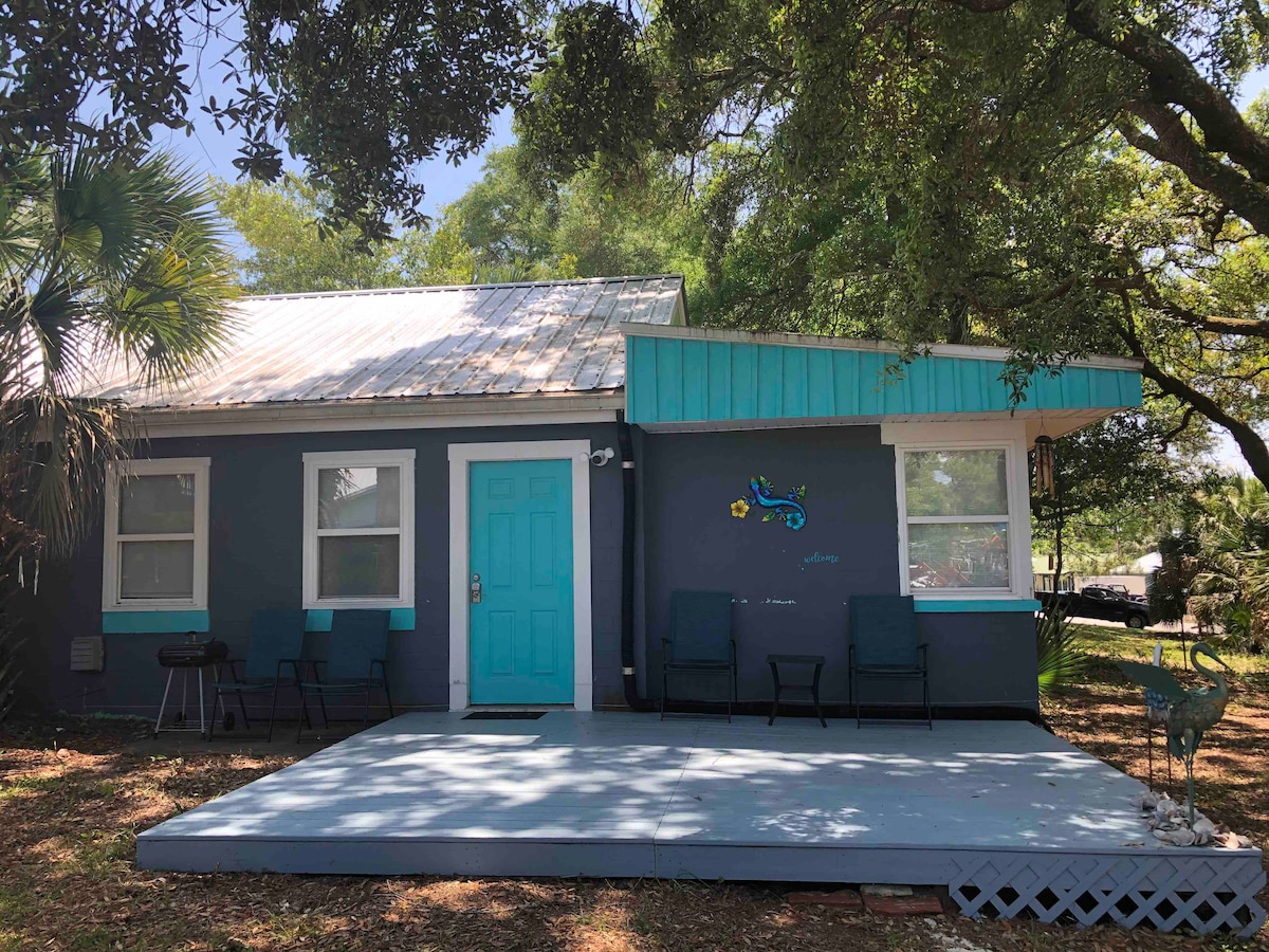 The cottage exterior features a shaded porch with two chairs and a vibrant blue door. A small area of outdoor flooring provides space for relaxation, while lush greenery surrounds the home, creating a serene and inviting entryway.