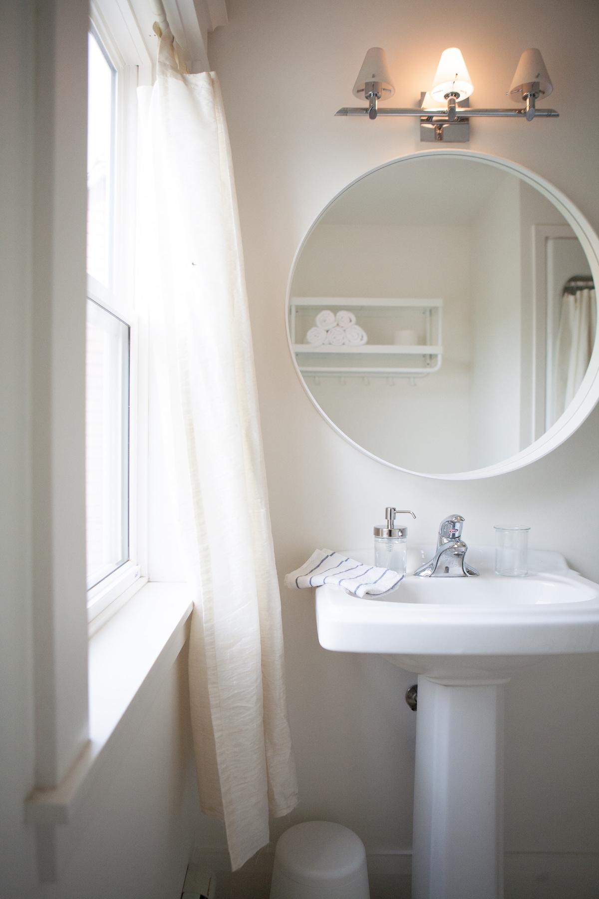 A bright bathroom features a pedestal sink with a modern faucet and a large round mirror above it. Natural light enters through a window covered by a light curtain. Shelves hold neatly stacked white towels, and a clear glass pitcher sits beside the sink.