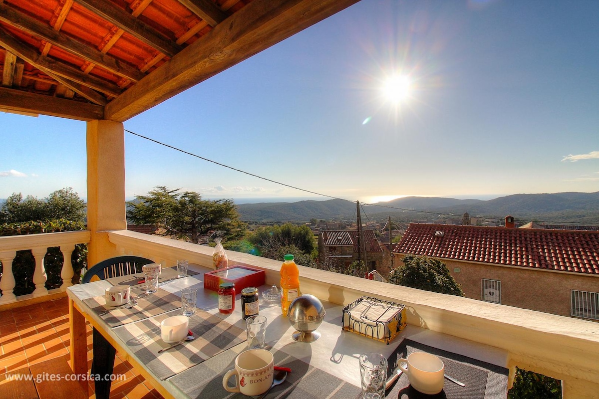 A dining area is situated on a terrace, featuring a table set with various items including drinks and utensils. The view showcases mountains and the sea with the sun shining brightly in the sky, creating a serene atmosphere. The space is framed by wooden beams.