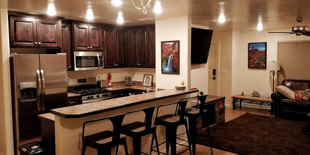 A modern kitchen space is showcased, featuring dark wood cabinetry and stainless steel appliances. A spacious countertop extends into a breakfast bar with four high-backed stools. Recessed lighting enhances the warm atmosphere, while artwork and a cozy seating area are visible in the background.