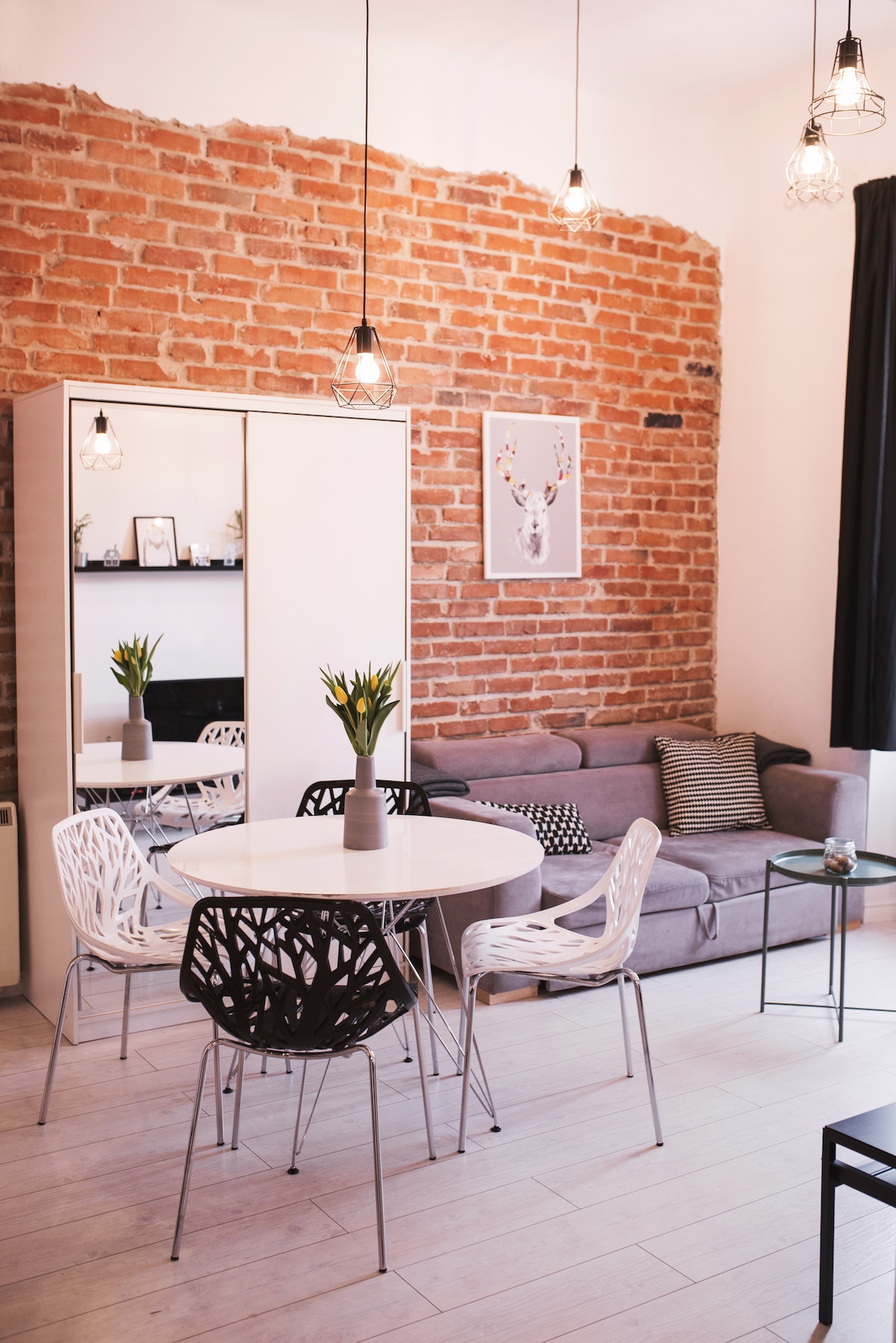 The living area features a blend of modern and rustic design, highlighted by an exposed brick wall. A round dining table with stylish chairs stands beside a cozy sofa, creating an inviting space. Pendant lighting adds a touch of warmth to the room.