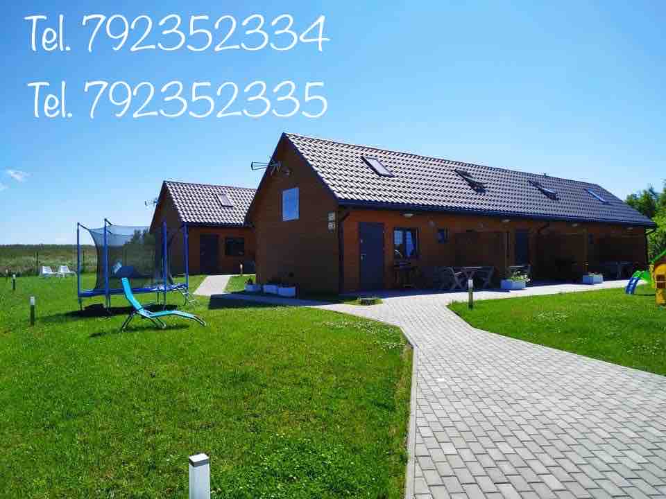Two quaint wooden cottages are set amidst a lush green lawn, accessible via a stone pathway. The cottages feature sloped roofs with skylights. Outdoor seating areas are visible, inviting relaxation in a peaceful environment, with a trampoline and lounge chair nearby.