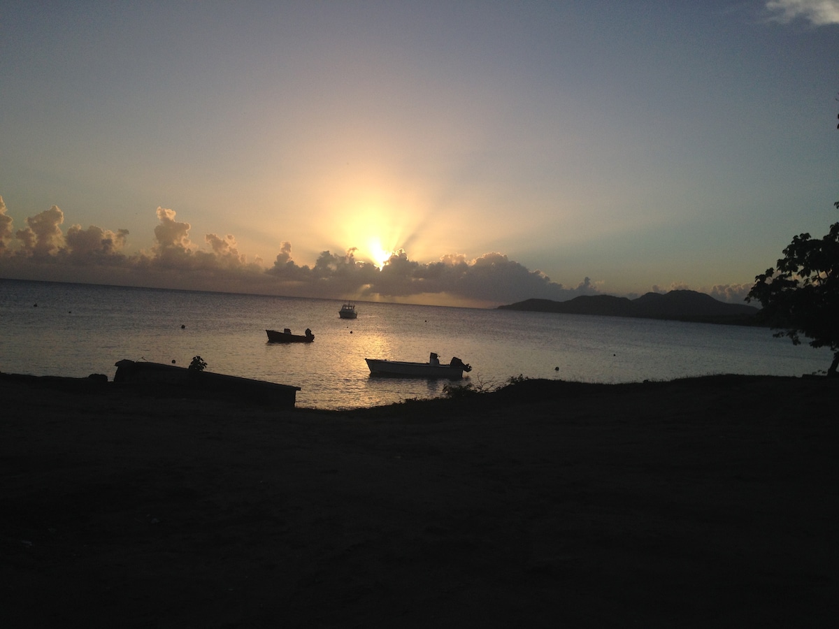 A serene beach scene captures the sun setting over the water, casting a warm glow across the horizon. Small boats are gently anchored in the foreground, while the coastline is framed by lush greenery, creating a tranquil atmosphere.