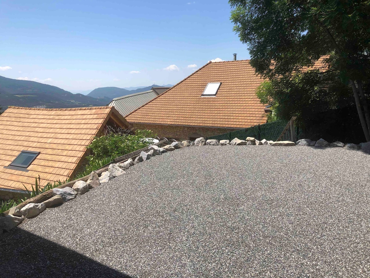 A gravel parking area is bordered by a decorative stone wall. In the background, terracotta rooftops and green hills are observed under a clear blue sky. Nearby vegetation provides a serene atmosphere, offering a glimpse of the surrounding landscape.