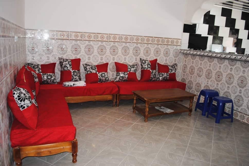 A bright seating area is presented with a collection of red couches adorned with black and white patterned cushions. A wooden coffee table occupies the center, complemented by a pair of blue stools. The walls feature intricate tile patterns, enhancing the overall aesthetic.