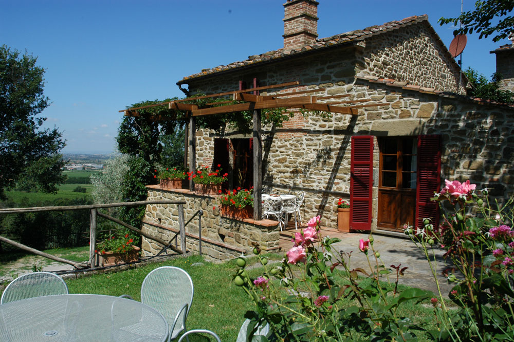 A rustic stone villa is set against a serene countryside backdrop, featuring charming wooden shutters and a welcoming entrance. A small outdoor dining area with light metal chairs is positioned near blooming rose bushes. The property is surrounded by lush greenery, enhancing the peaceful atmosphere.