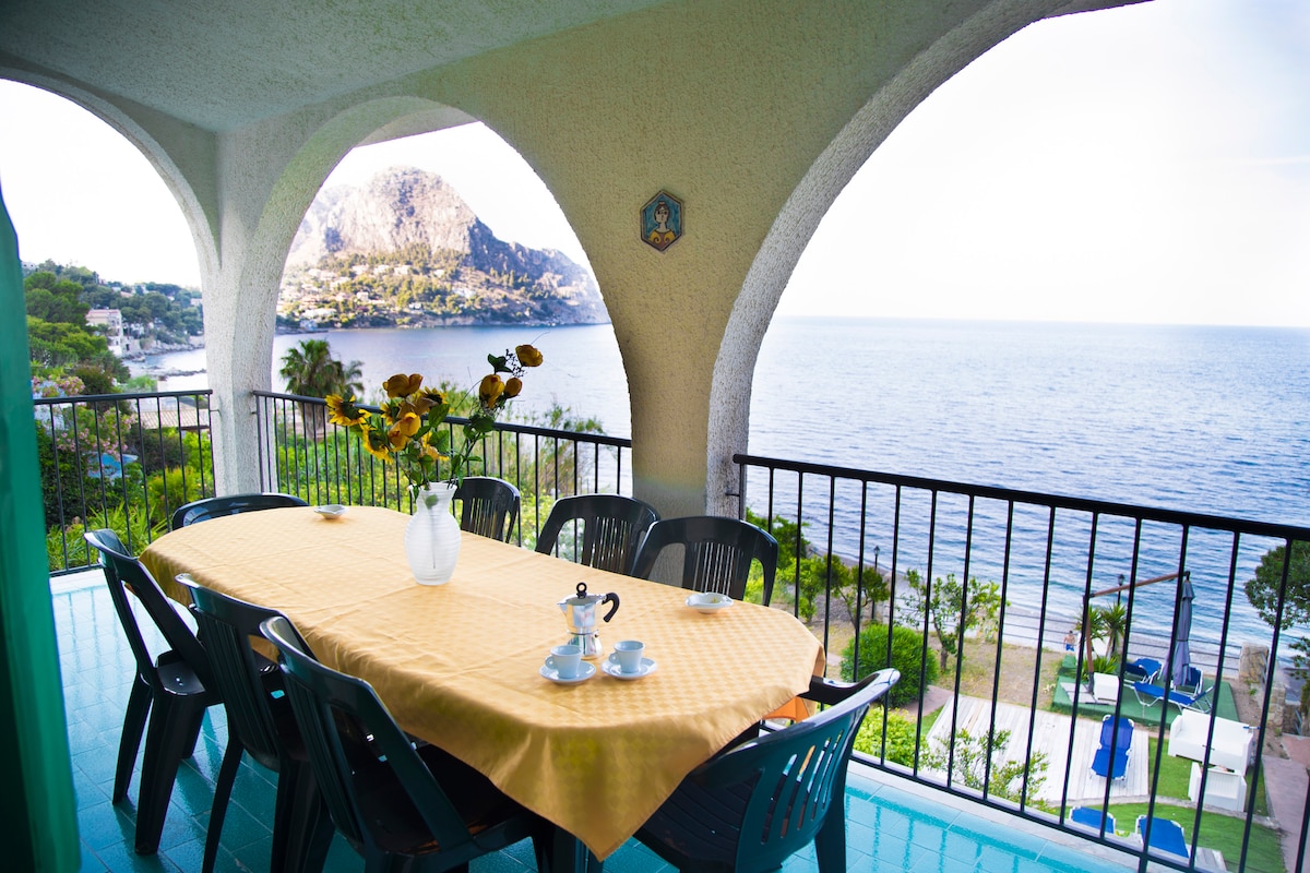 A spacious outdoor dining area is situated on a balcony, featuring a large table covered with a yellow tablecloth. Eight black chairs are positioned around the table. A vase with sunflowers is placed at the center, while the stunning view of the sea and mountains is visible in the background.