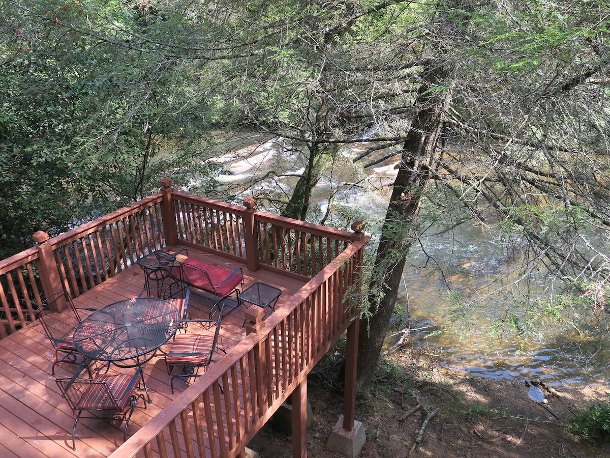 An outdoor deck is seen overlooking a flowing creek, surrounded by trees. The deck is equipped with metal furniture, including a circular table and several chairs. The gentle sound of the water enhances the natural ambiance of the setting.