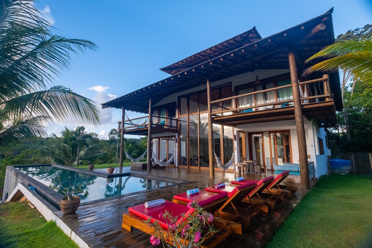 The exterior of the house features a modern two-story design, with large glass doors opening onto a spacious deck. An infinity pool lines the edge, surrounded by sun loungers and tropical plants, providing a serene atmosphere with lush greenery in the background.