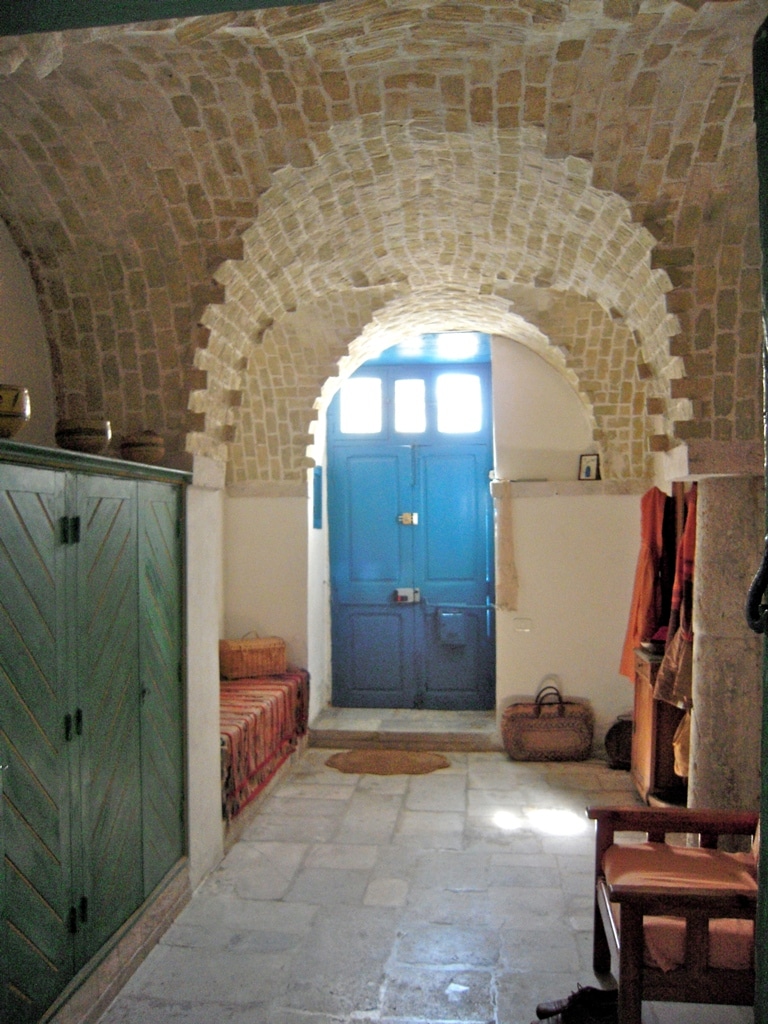 A traditional hallway is characterized by high, arched brick ceilings and a blue wooden door that provides an entry point. The space features wooden storage units against the walls and simple furnishings, creating an inviting entrance that reflects local architectural styles.