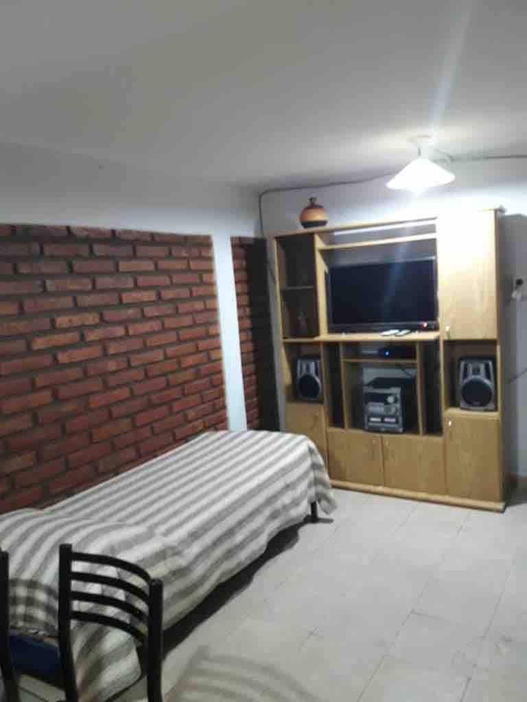 A modest living area features a single bed covered with a striped blanket, a wooden cabinet housing a television and speakers, and exposed brick walls. A lamp illuminates the space from the ceiling, creating a functional environment.