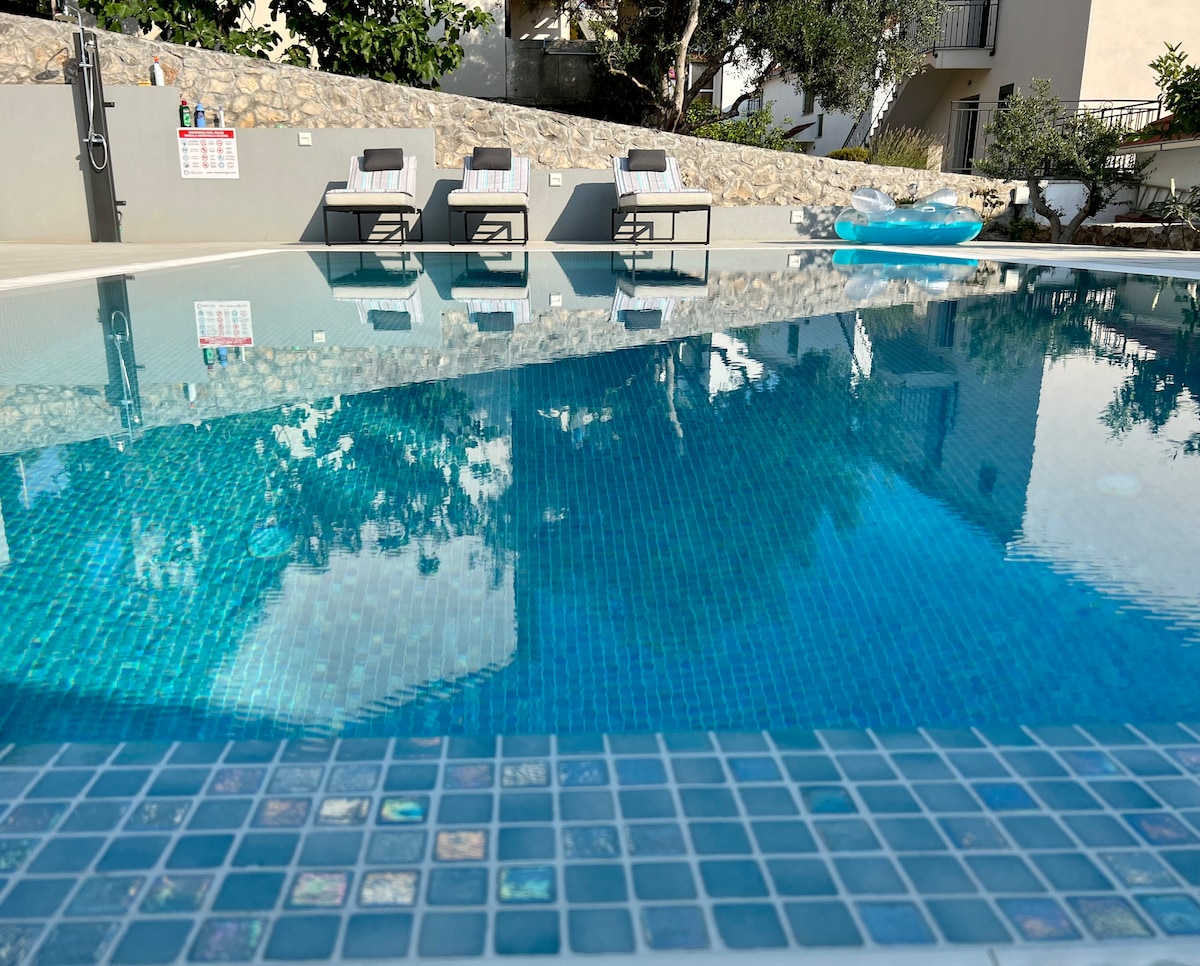 A serene outdoor space features a clear, inviting pool with a tiled edge. Three sun loungers are positioned along the water, surrounded by greenery and a stone wall, providing a tranquil atmosphere for relaxation.