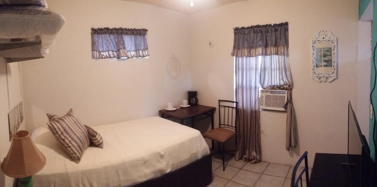 The room features a cozy setup with a spacious bed adorned with striped pillows. A small table and chair are positioned beside a window. An air conditioning unit and a television are visible. Natural light filters through sheer curtains, enhancing the simplicity of the space.