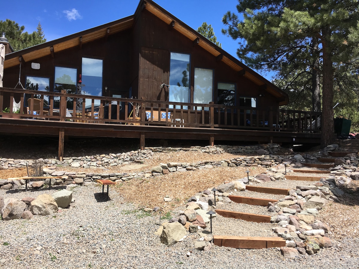 The exterior of a charming wooden home is showcased, featuring a spacious deck with multiple large windows that allow natural light to fill the interior. Stone pathways lead up to the deck, surrounded by landscaped gravel and natural elements, reflecting a peaceful outdoor environment.