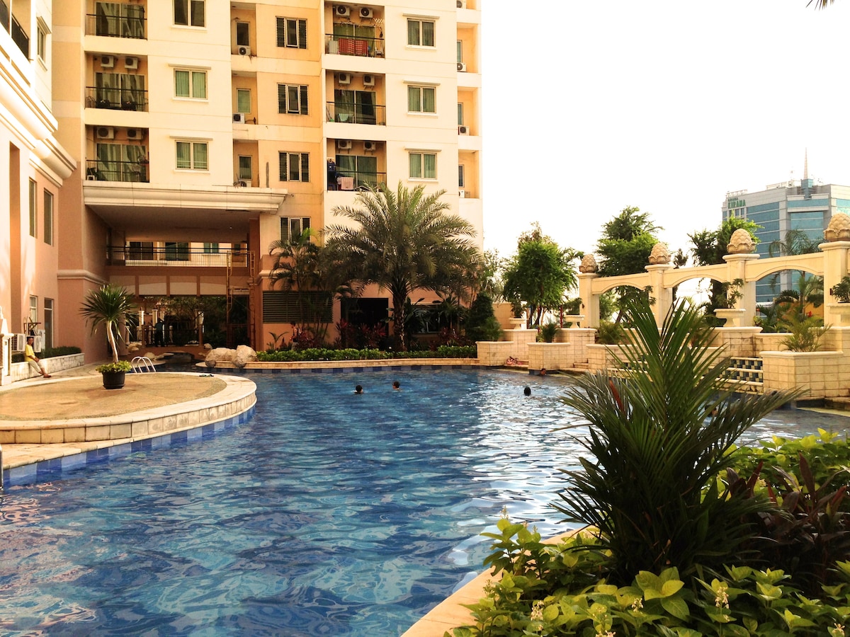 A large swimming pool is surrounded by lush greenery and palm trees, reflecting the clear blue sky. The building's facade features balconies and windows, with a peaceful atmosphere creating a welcoming environment for relaxation.