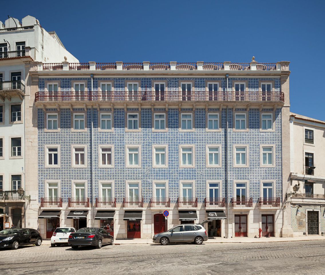 The building features a distinctive blue façade adorned with intricate tile patterns. Large windows are complemented by wrought-iron railings on the balconies. The entrance is framed by awnings, with a row of parked cars visible in front. Clear blue skies create a bright backdrop.