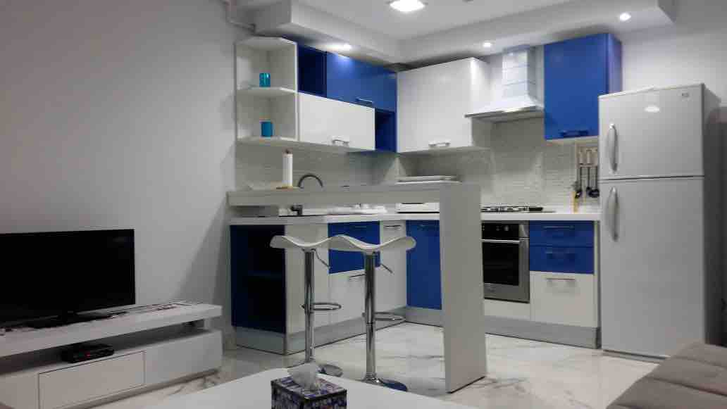 A modern kitchen features blue and white cabinetry with sleek countertops. A refrigerator stands to the right, while a gas stove is positioned within easy reach. Two modern bar stools are placed at a small, functional dining area. Light fixtures above illuminate the space.