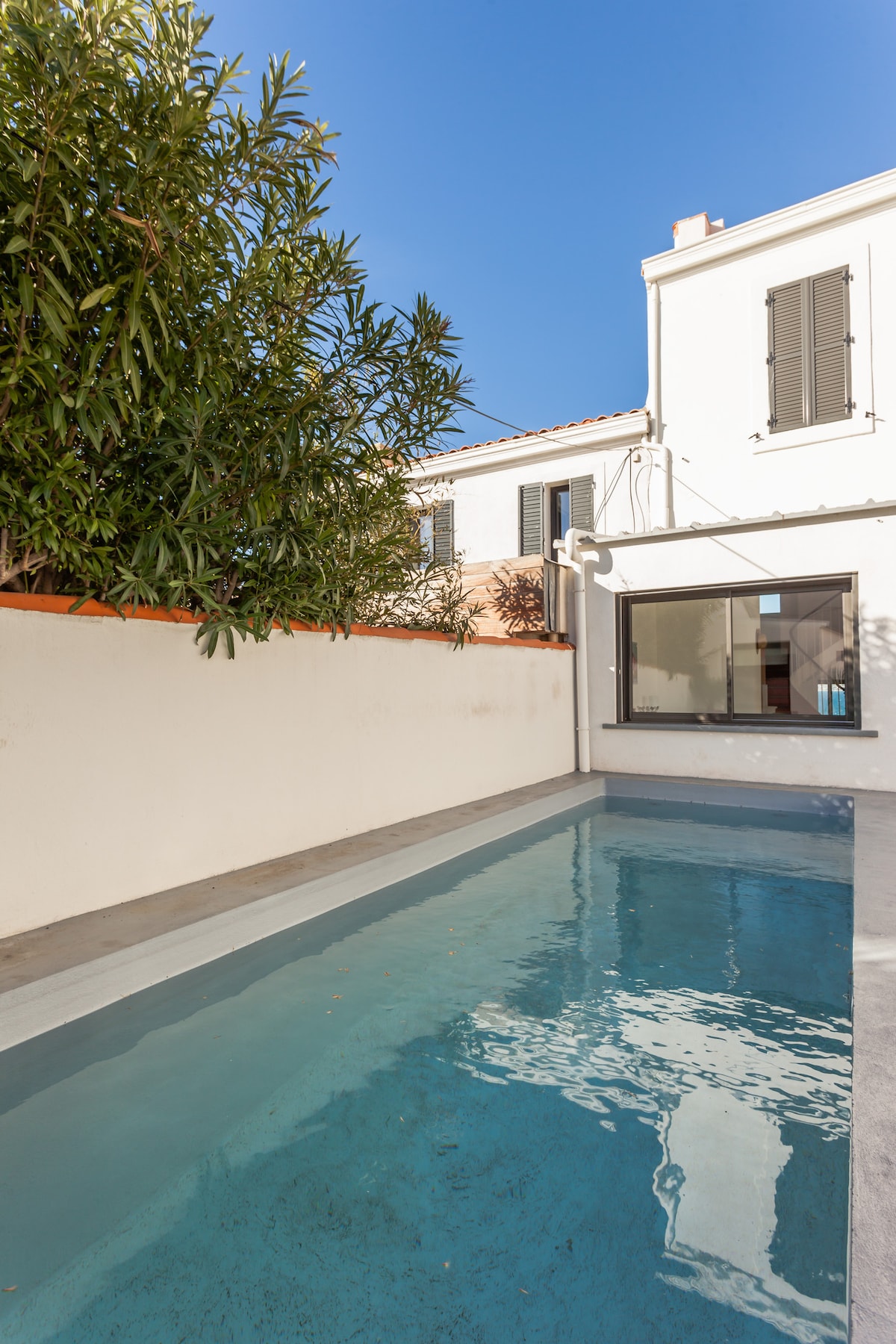 A rectangular swimming pool with clear blue water is situated within a private courtyard. Surrounded by a smooth gray pool deck, the area is enclosed by a low white wall and features greenery on one side, allowing for a serene atmosphere under clear skies.