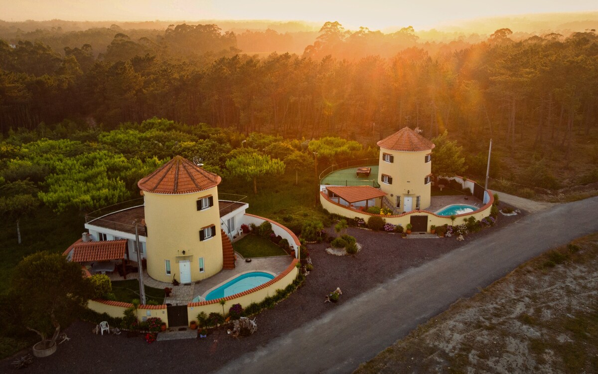 Two round structures with distinctive terracotta roofs are visible, surrounded by lush greenery. Each building features a private pool and outdoor space, highlighted by the warm glow of sunset. A gravel pathway runs alongside, leading to a serene environment.