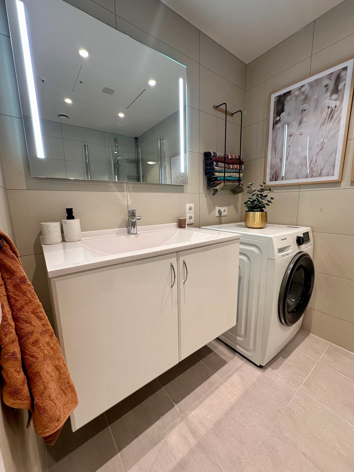 A modern bathroom features a sleek white vanity with a sink and a mirror above it, complemented by a decorative wall art piece. A washing machine sits beside the vanity, with a towel hanging on a nearby rack. Soft lighting provides a bright ambiance.