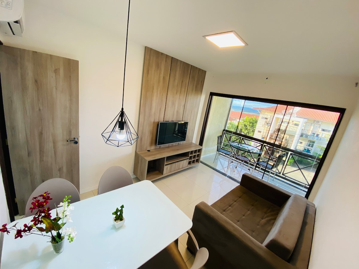 A modern living area features a cozy brown sofa positioned next to a television mounted on a stylish wooden panel. A dining table with chairs is set beside a large window, offering natural light and a view of the surroundings.