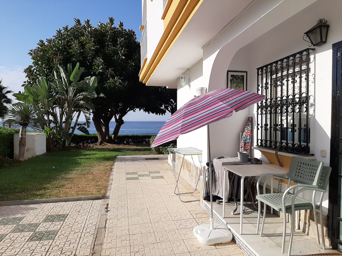 A patio area is visible, featuring a table with two chairs and a striped umbrella providing shade. Lush greenery is situated nearby, with a view of the ocean in the background. The paved pathway leads toward the beach, enhancing outdoor accessibility.