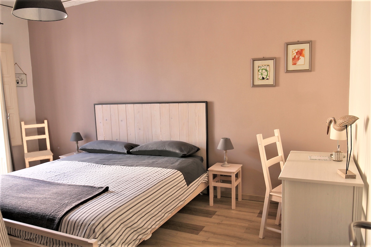 A private bedroom features a spacious bed with neutral-colored bedding. Two simple bedside tables with lamps are positioned beside the bed. A small desk with a lamp is located near the wall, and framed artwork adorns the walls, adding a touch of decor.