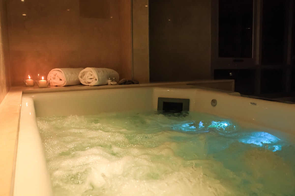A relaxing hot tub is illuminated by soft blue lighting, surrounded by candles. Neatly rolled towels rest on the nearby surface, contributing to a serene atmosphere. The bubbling water creates an inviting setting for relaxation.