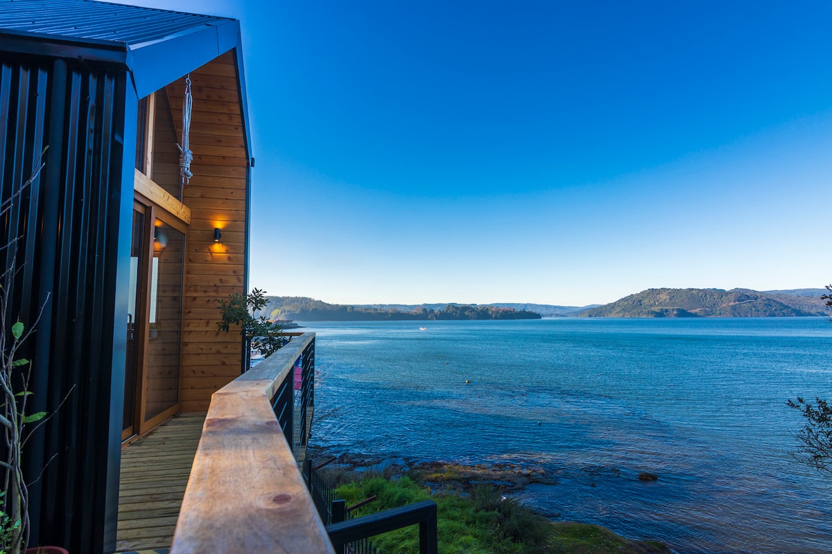 A wooden deck extends along the exterior, offering panoramic views of the calm water and distant hills. The clear blue sky complements the serene landscape, inviting moments of tranquility amidst nature.