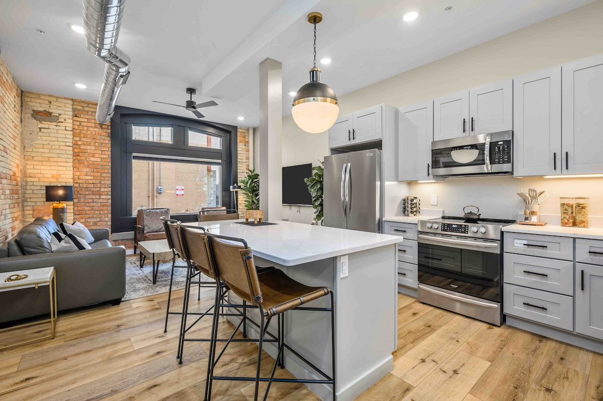 An open-concept kitchen and living area features modern stainless steel appliances and a spacious quartz countertop island with seating for three. Exposed brick walls add character, while large windows provide ample natural light. Stylish furnishings complement the space, promoting a welcoming ambiance.