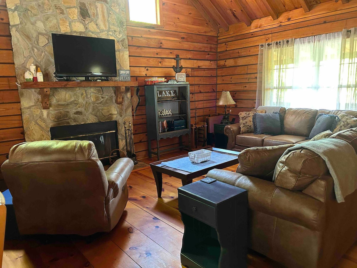 A cozy living area is presented with a stone fireplace as the focal point. Leather couches and an armchair provide ample seating. A TV is mounted above the fireplace, and a wooden coffee table is positioned centrally. Natural light filters through sheer curtains.