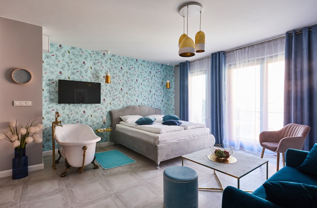 A spacious bedroom features a large, upholstered bed with decorative pillows, complemented by a vintage-style bathtub. The room is adorned with floral-patterned wallpaper and is illuminated by natural light through floor-to-ceiling windows, flanked by soft blue curtains. A cozy armchair and a small coffee table complete the setting.