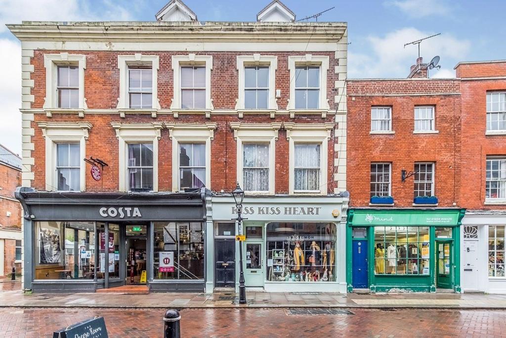The historic building features a classic red brick facade, showcasing three levels with large windows. A Costa coffee shop occupies the ground floor, flanked by a boutique and a green-fronted shop. The street is paved and lined with street lamps, enhancing the charm of the area.