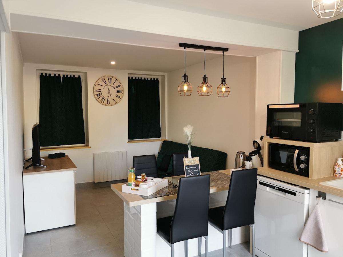 A modern kitchen and living area are depicted, featuring a dining table with four black chairs. A comfortable green sofa is positioned nearby, complemented by dark curtains over the windows. A wall clock and various kitchen appliances, including a microwave and coffee maker, are visible.