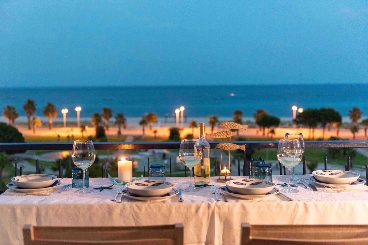 An outdoor dining table is set for a meal, overlooking a beach illuminated by evening lights. Glasses and plates are arranged neatly, with a candle providing soft illumination. A gentle sea breeze can be felt as the tranquil ocean waves lap against the shore.