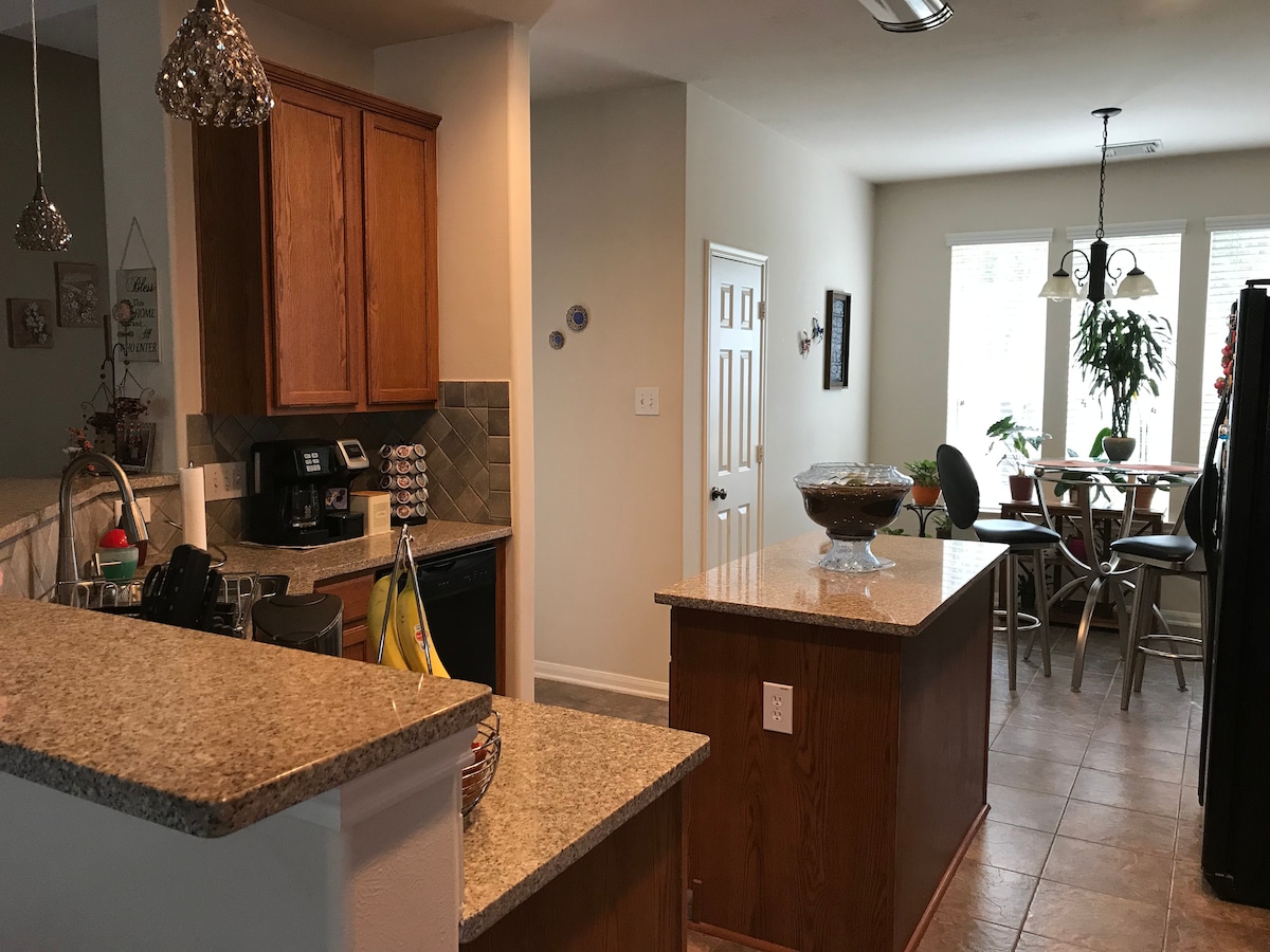 The kitchen features warm wood cabinetry with granite countertops, complemented by stainless steel appliances. An island offers additional preparation space, while a dining area with a table and chairs is visible through the open layout. Natural light enters through large windows.