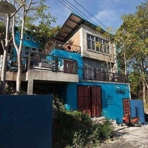 The three-story building showcases a unique loft style with a striking blue exterior. Large windows and a spacious balcony create a bright atmosphere, while surrounding trees offer natural shade. The landscape is characterized by gentle slopes and greenery, contributing to the serene setting.