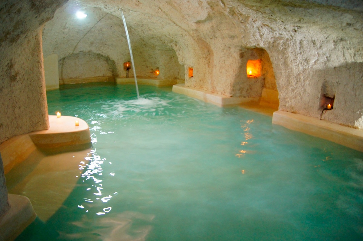 A serene underground natural cave is depicted, featuring a warm, salt-water pool. Soft lighting from the embedded candles illuminates the rugged stone walls, creating a calming atmosphere. Water gently flows from a narrow channel, enhancing the tranquil setting.